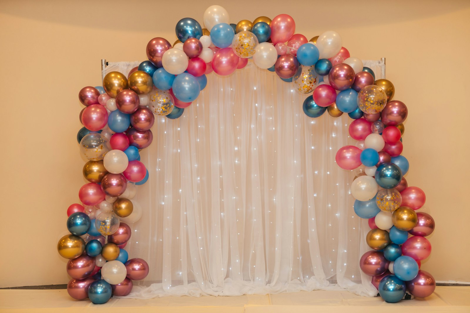 Rose-gold, blush and blue balloon arch framing a sheer curtain with fairy lights
