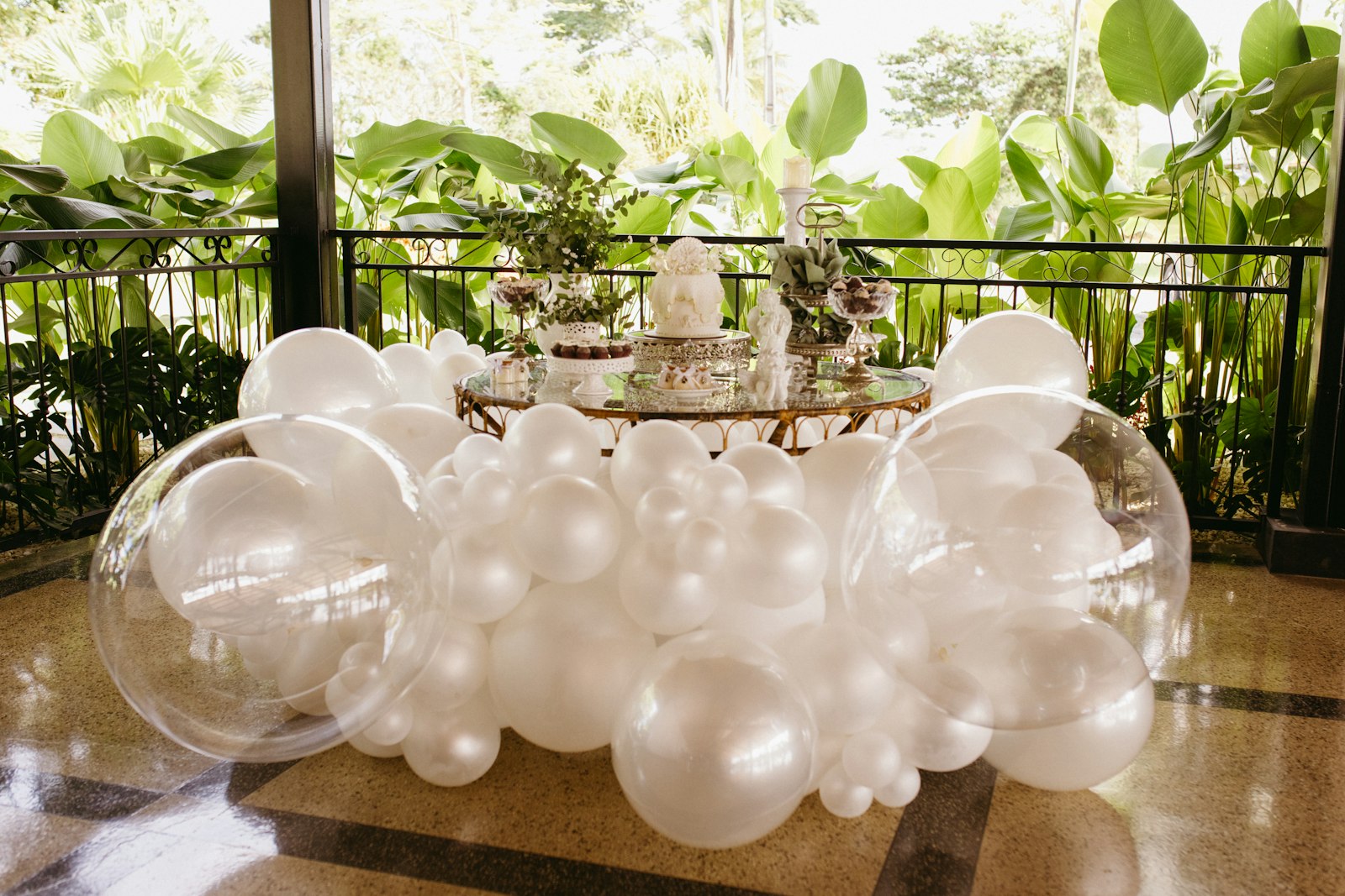 Cloud of pearl-white and clear balloons around a gold mirrored dessert table with a white cake