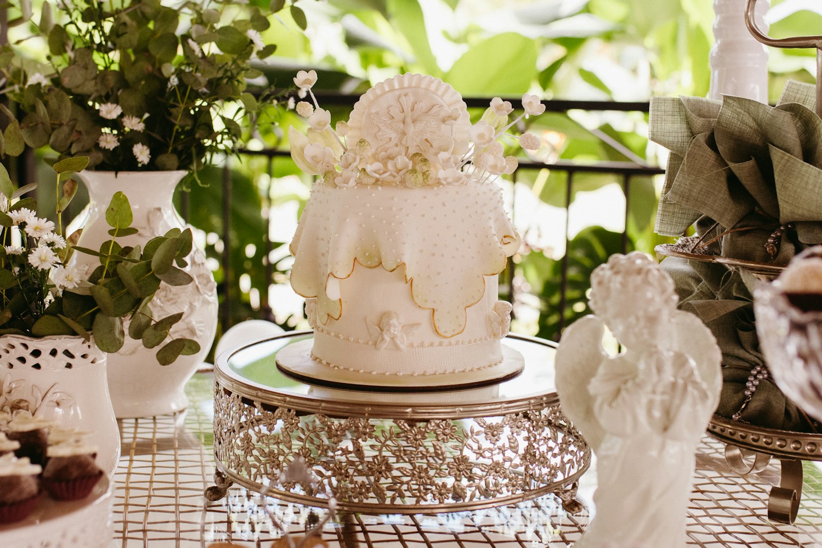 White lace-detailed cake on a silver filigree stand with an angel figurine and white flowers