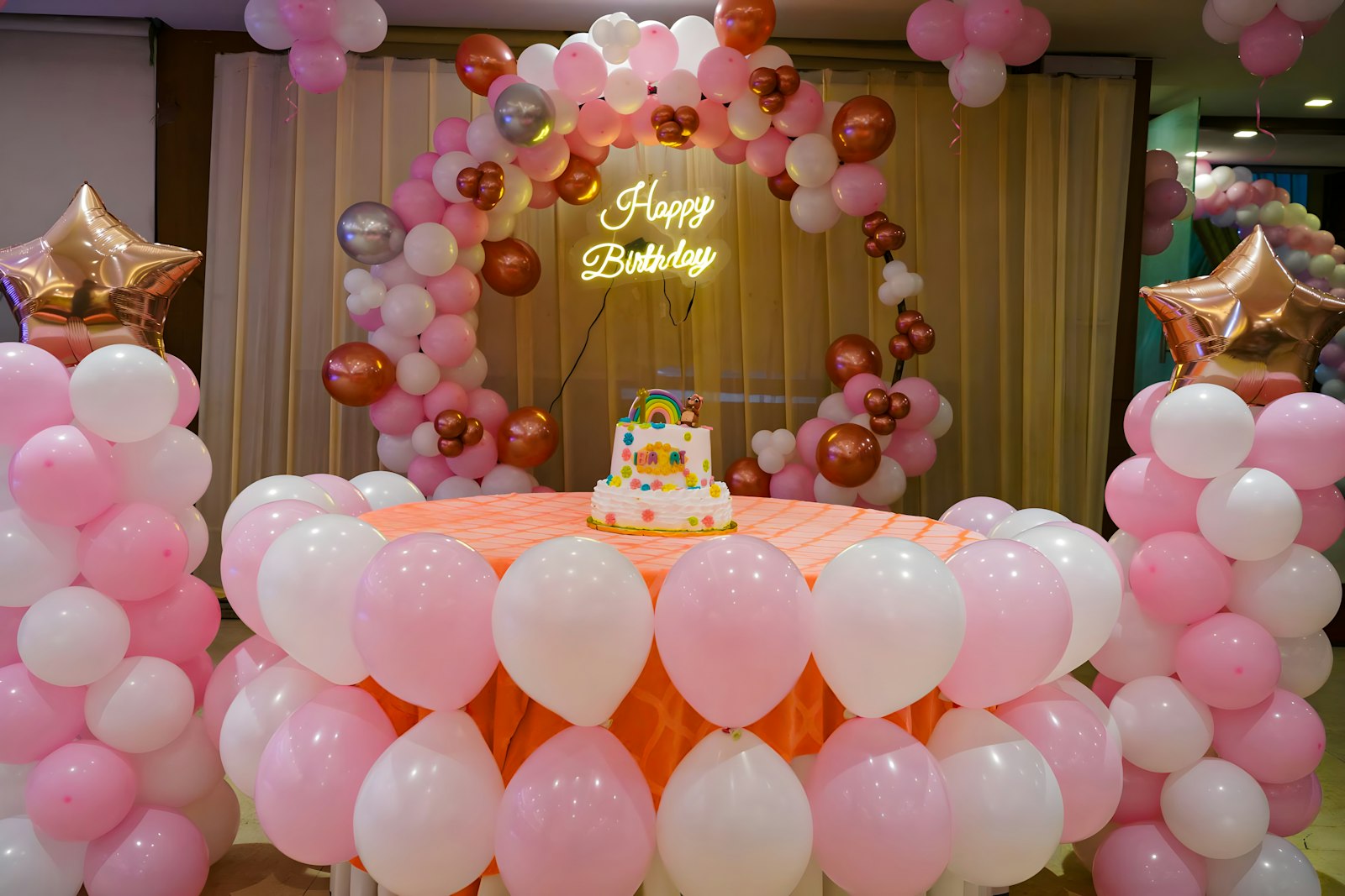 Pink, white and rose-gold balloon arch and columns around a cake table with a neon Happy Birthday sign