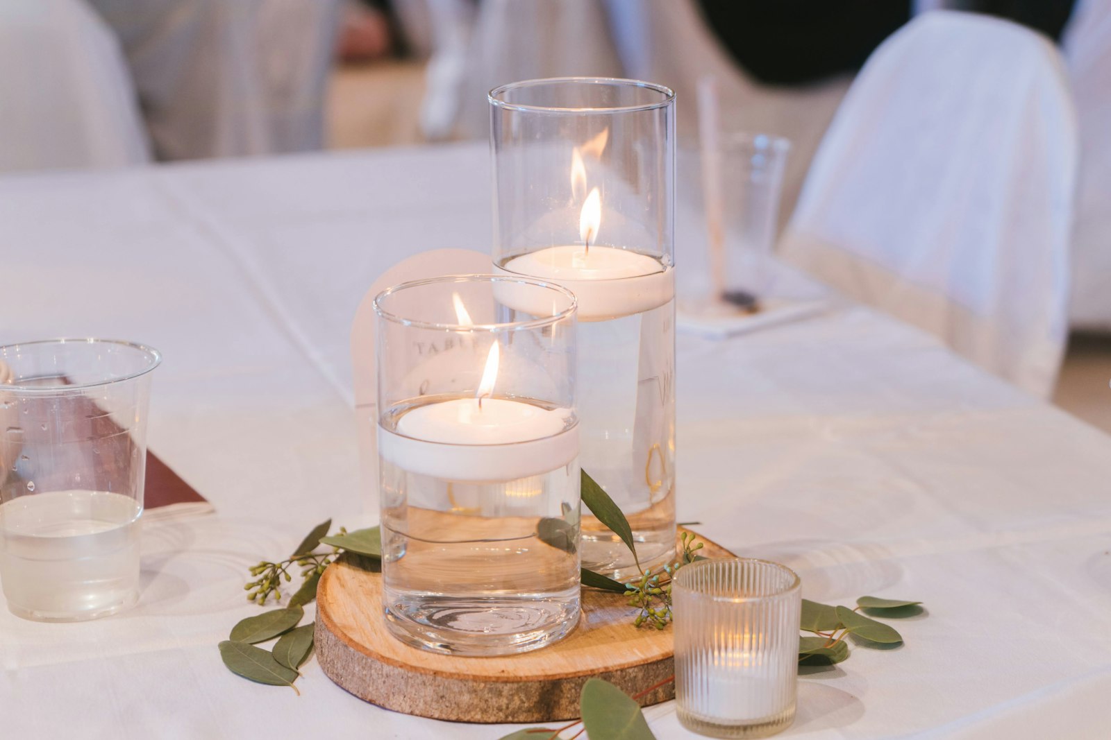 Low centrepiece of floating candles in two glass cylinders on a wood slice, with eucalyptus sprigs and a ribbed votive on white linen