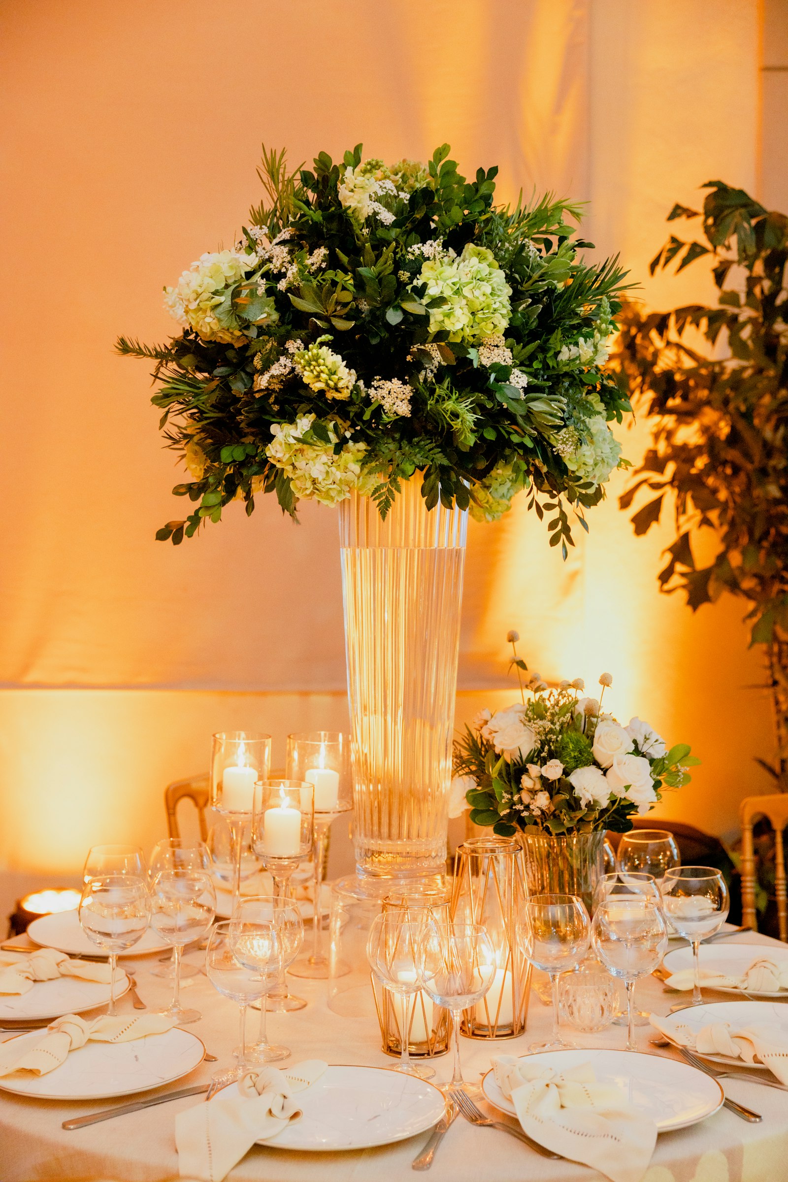 Tall fluted glass vase of green hydrangea and greenery above pillar candles and gold geometric lanterns, with warm amber uplighting on draped walls