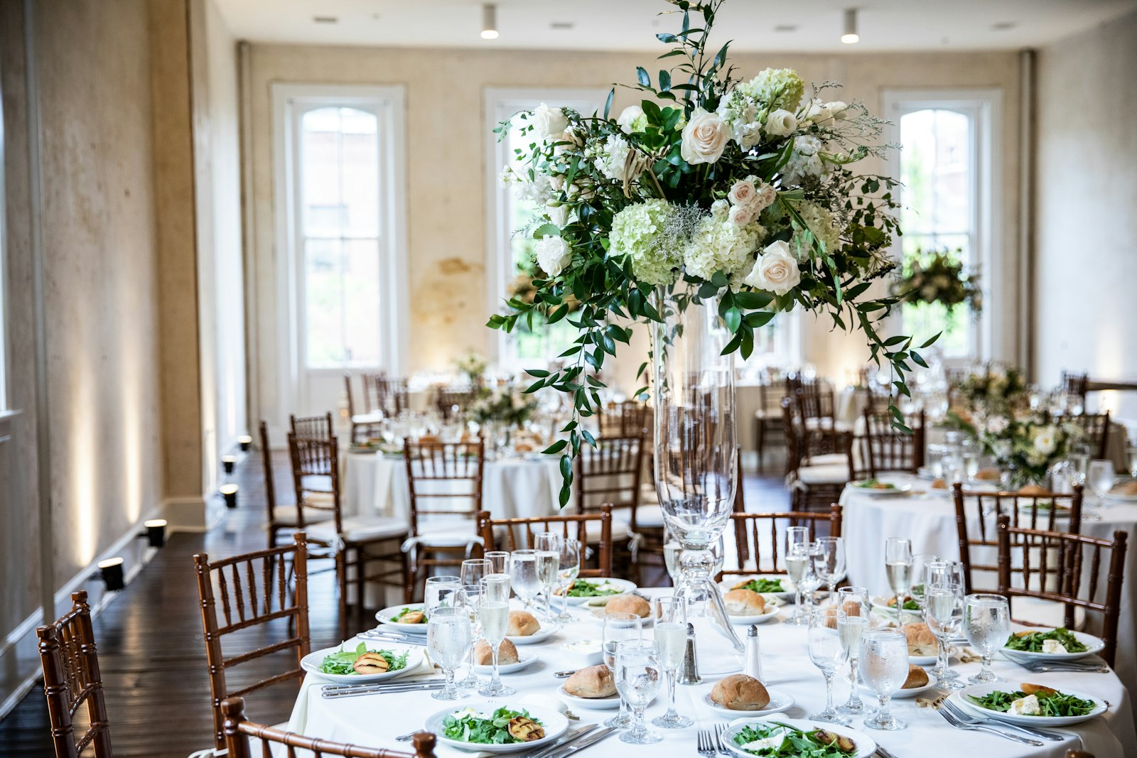 Tall glass vase of white roses, green hydrangea and trailing greenery on a round reception table with wooden Chiavari chairs