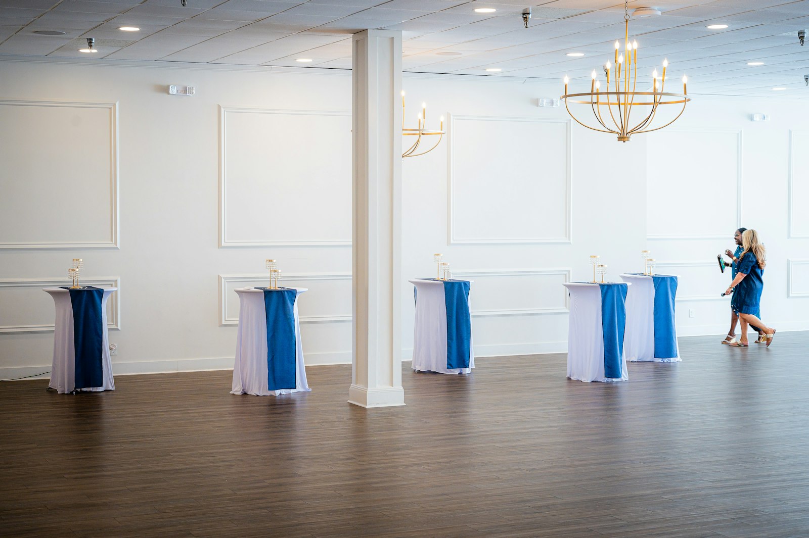 Bright event room with white and cobalt-blue spandex cocktail tables under gold chandeliers