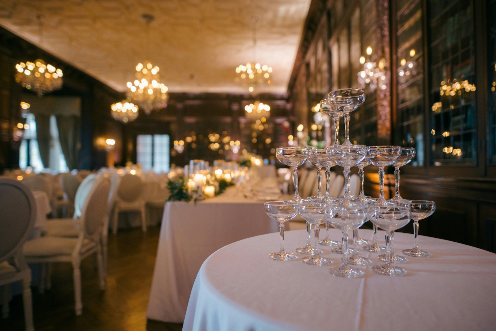 Champagne coupe tower on a white-linen table in a dining room lit by chandeliers