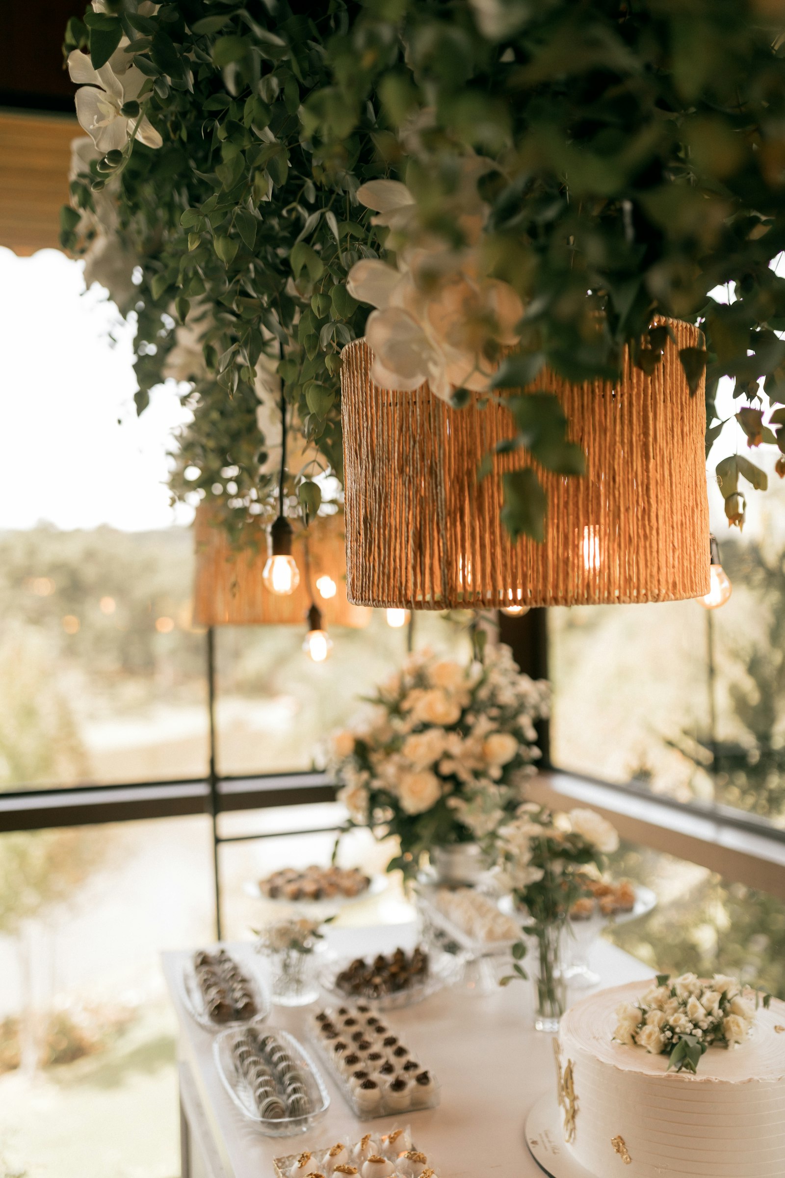 Rattan pendant lights draped in greenery above a white dessert table with cream flowers