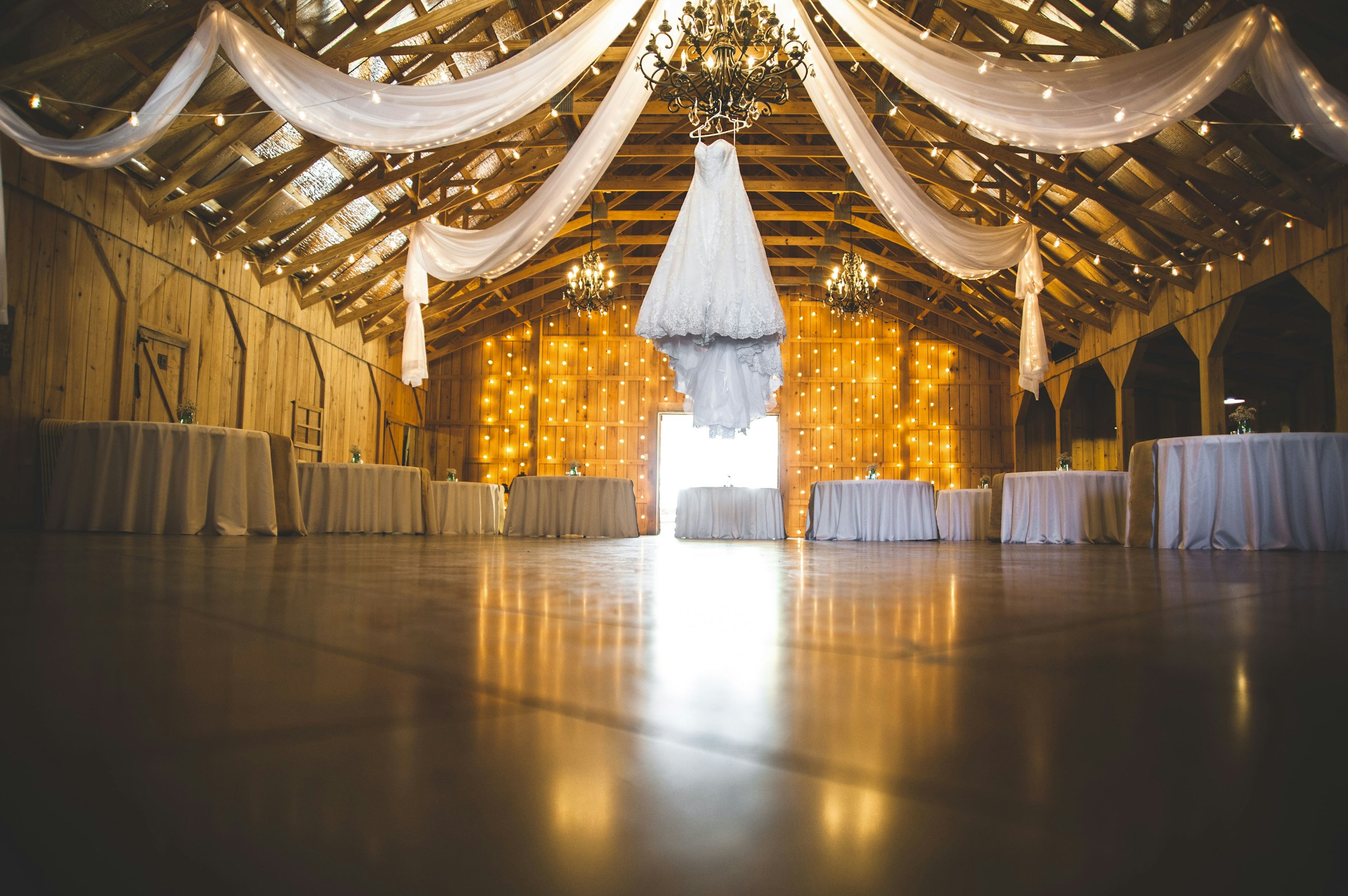 Barn reception with white ceiling swags, crystal chandeliers and a wall of string lights
