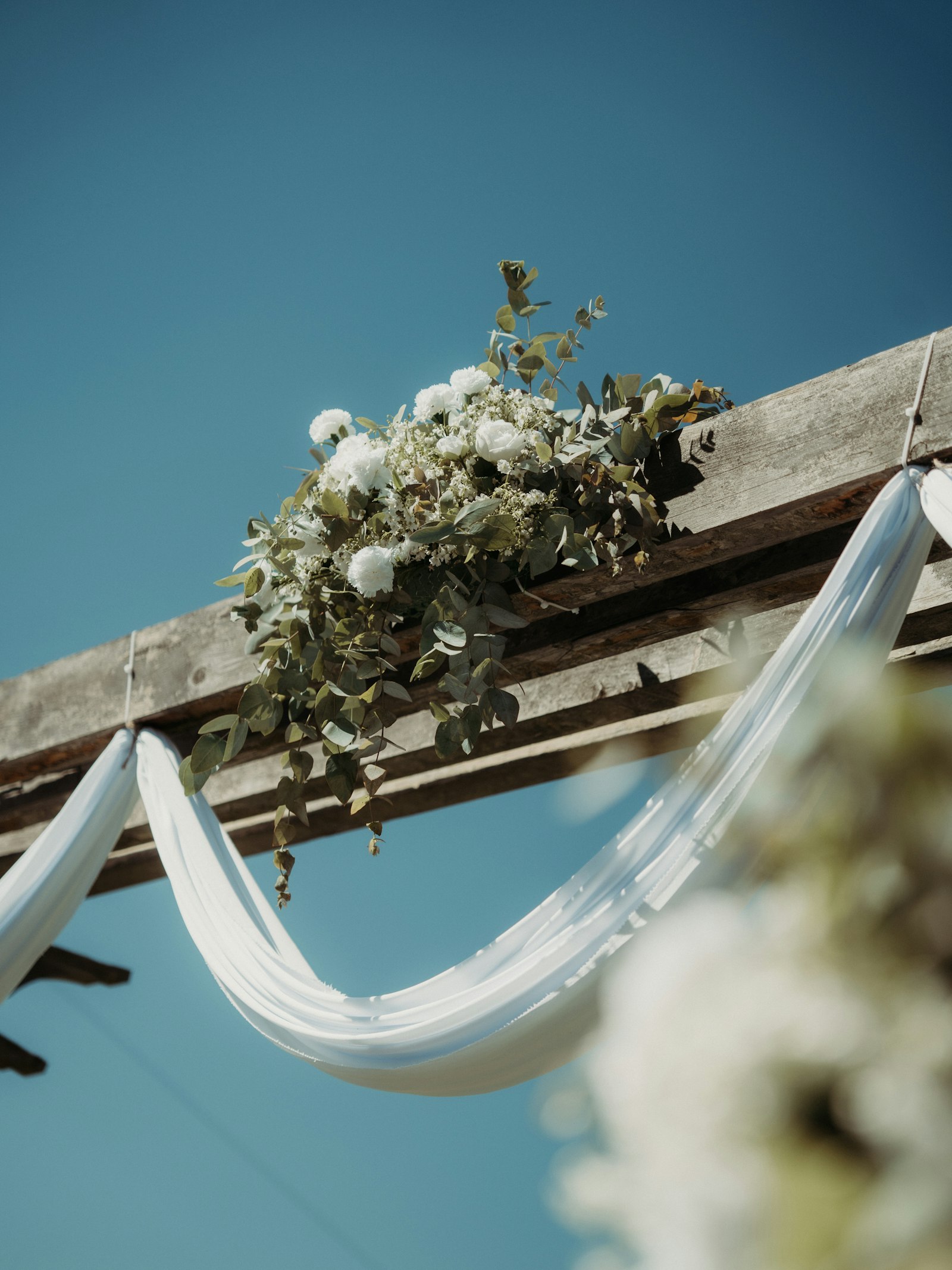 Rustic wooden arbour topped with white roses, carnations, baby's breath and eucalyptus, with a white fabric swag against a clear blue sky