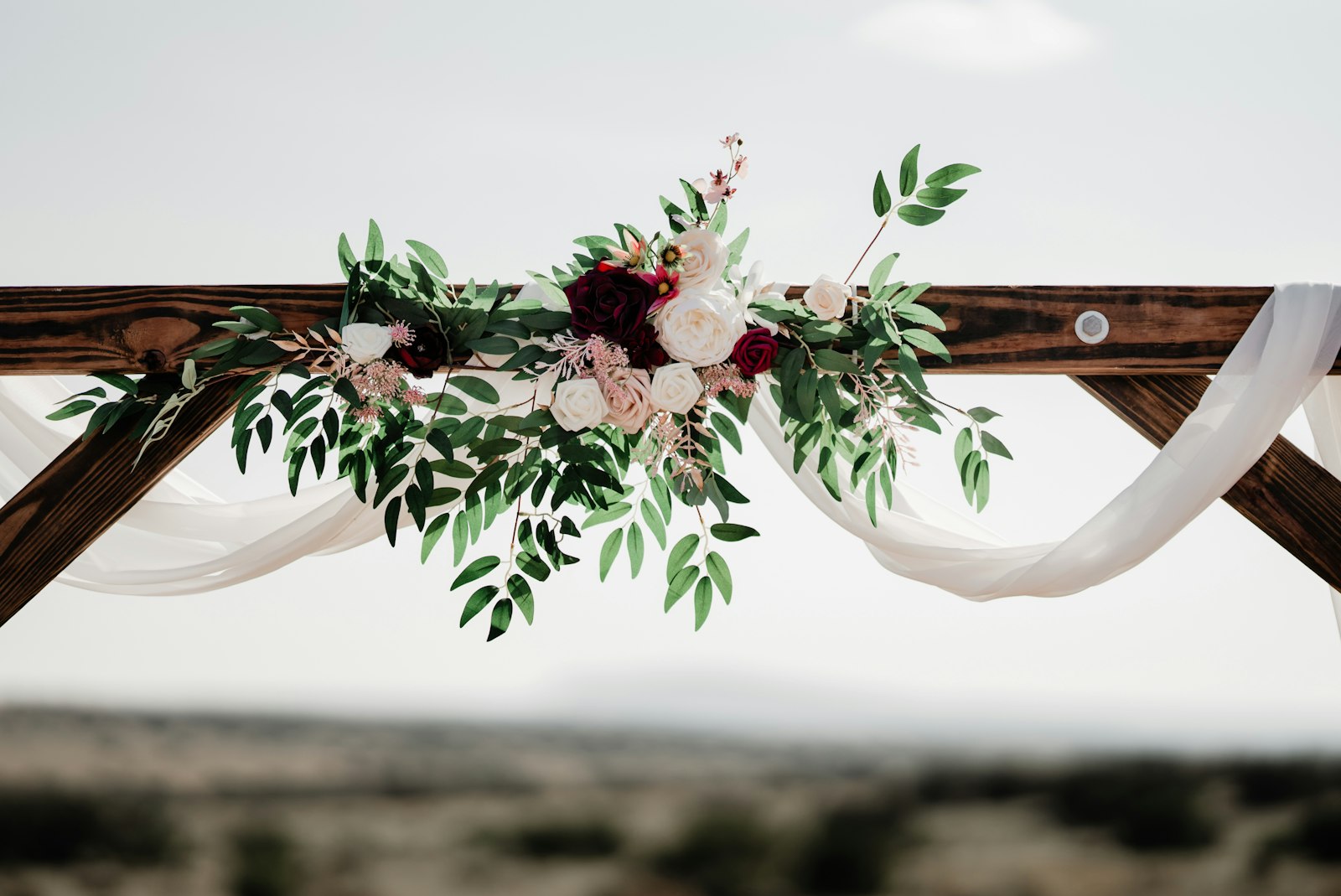 Wooden ceremony arch with a white chiffon swag and burgundy, blush and white roses with eucalyptus