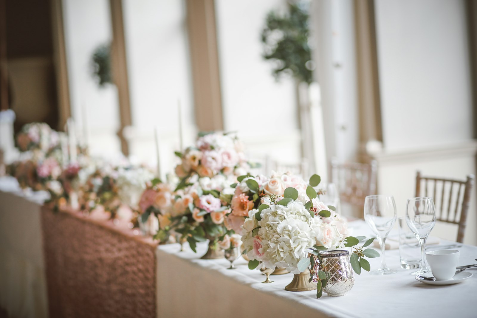 Head table lined with low gold compotes of blush and peach roses, white hydrangea and eucalyptus over a rose-gold sequin runner