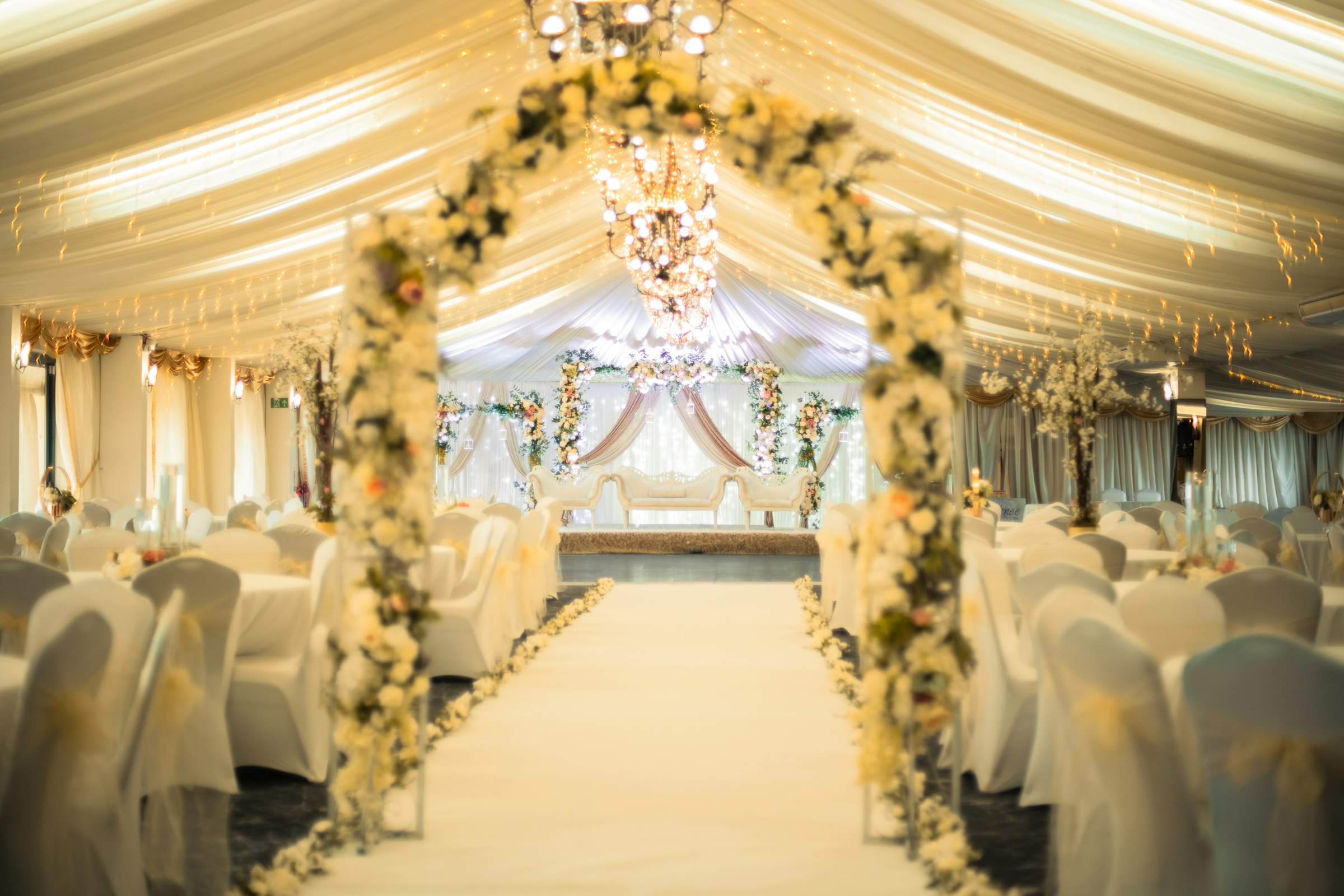 Reception tent with an ivory draped ceiling, crystal chandeliers and a white floral archway over a white aisle runner leading to the stage