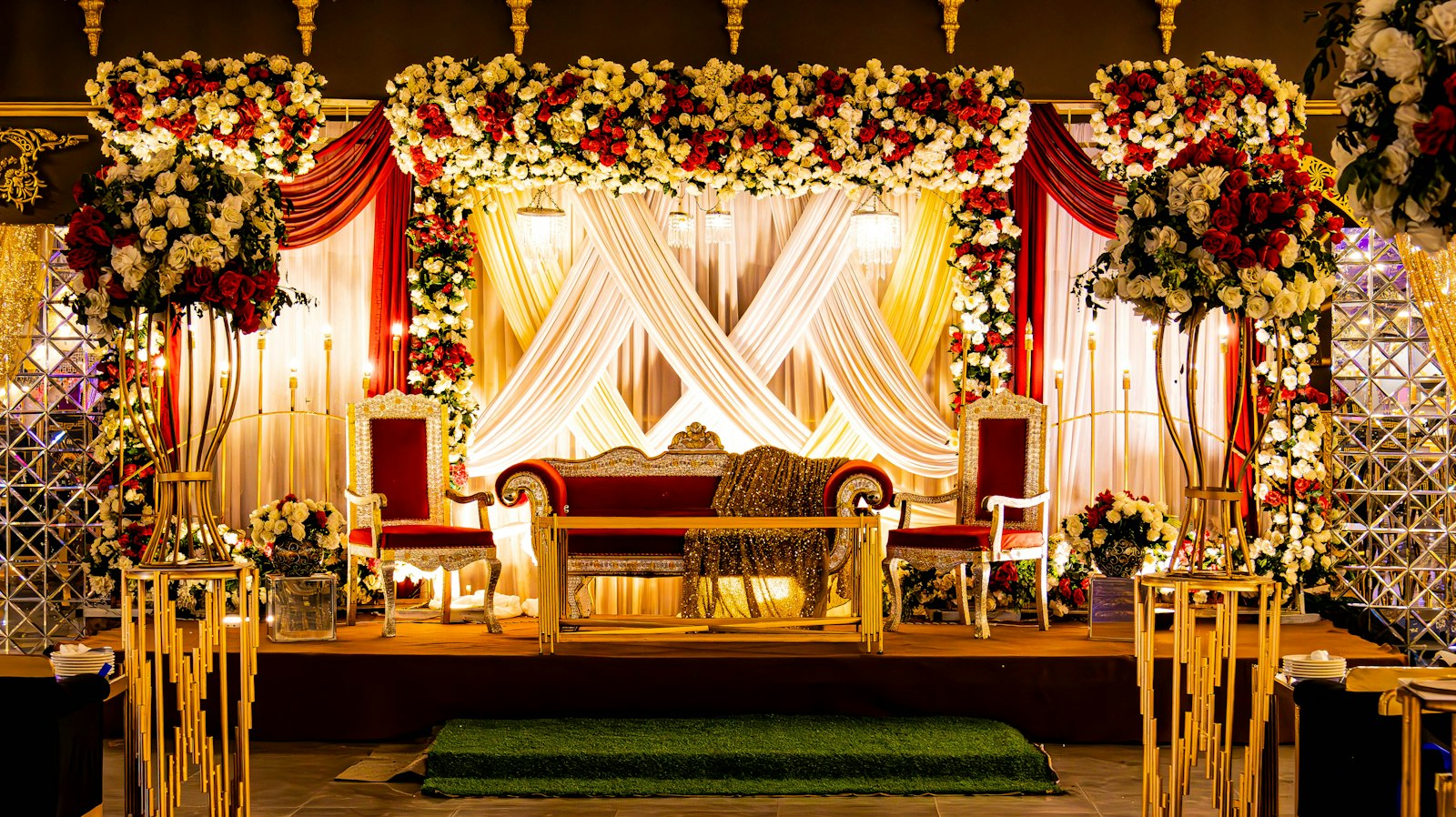 Reception stage with crossed ivory and gold drapes, a canopy of red and white roses, a red velvet loveseat between two carved chairs and tall gold floral stands