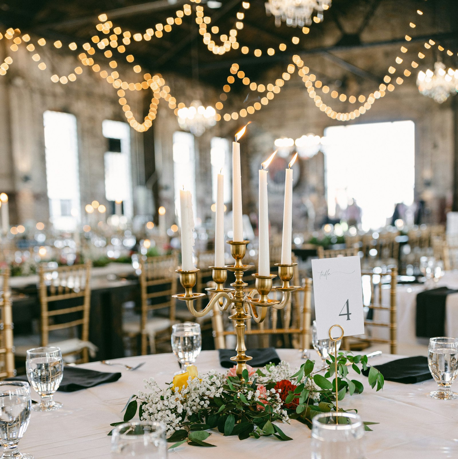 Round reception table with a gold five-arm candelabra and white taper candles rising from a ring of baby's breath and greenery, under string lights and crystal chandeliers